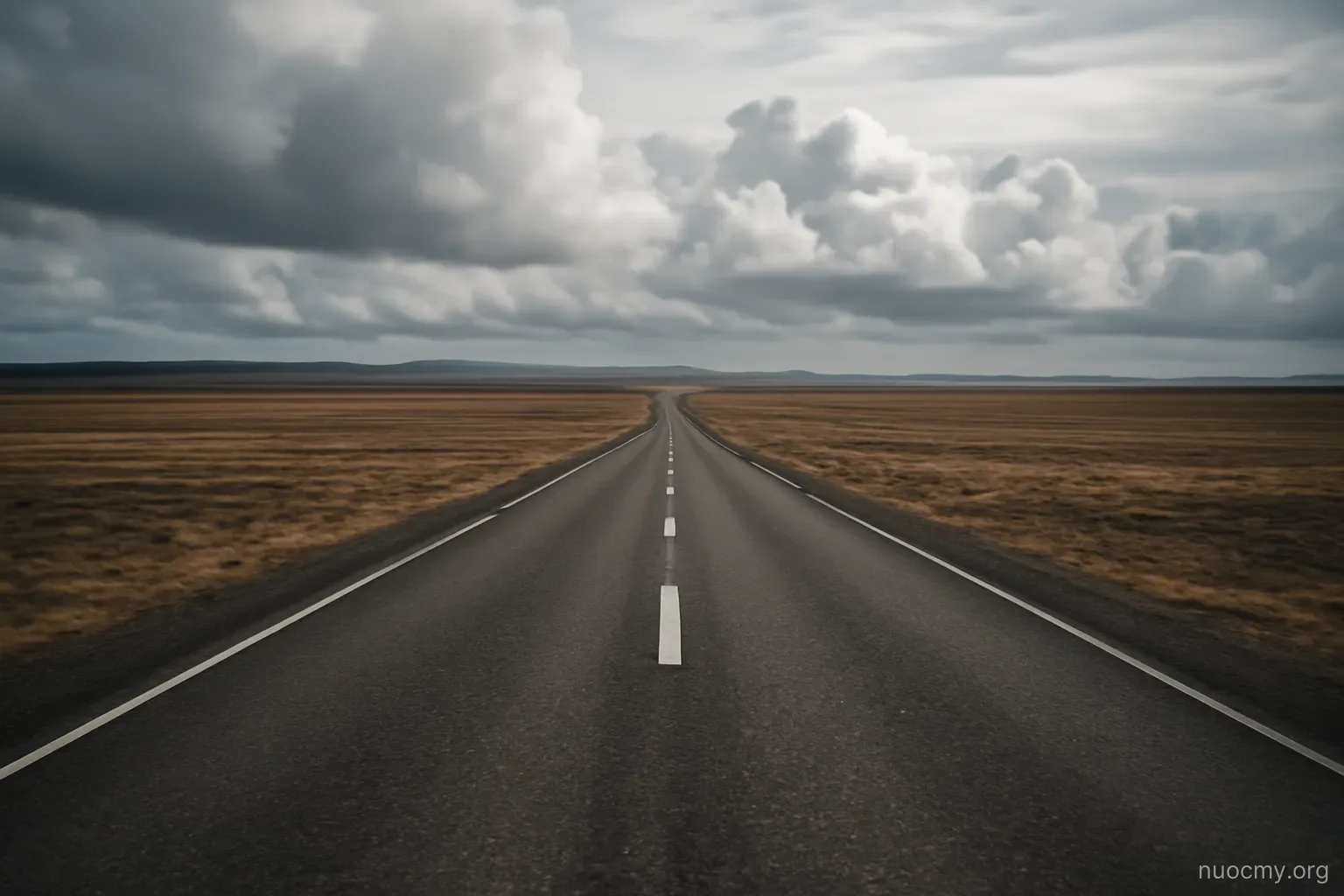 a bare landscape with distant clouds gathering over flat ground, a single road cutting through