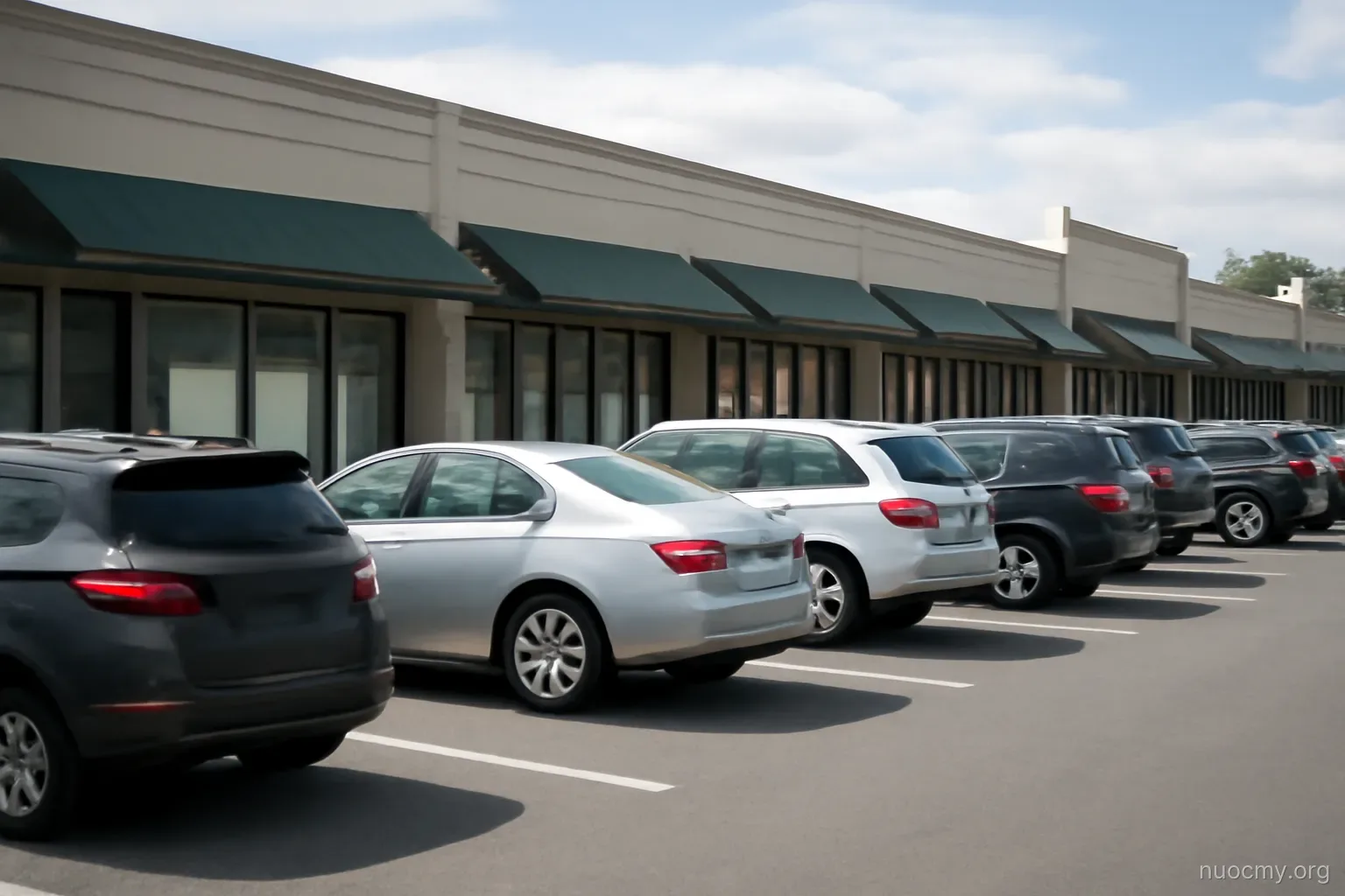 a strip mall parking lot with awnings over small shopfronts, cars parked in even rows